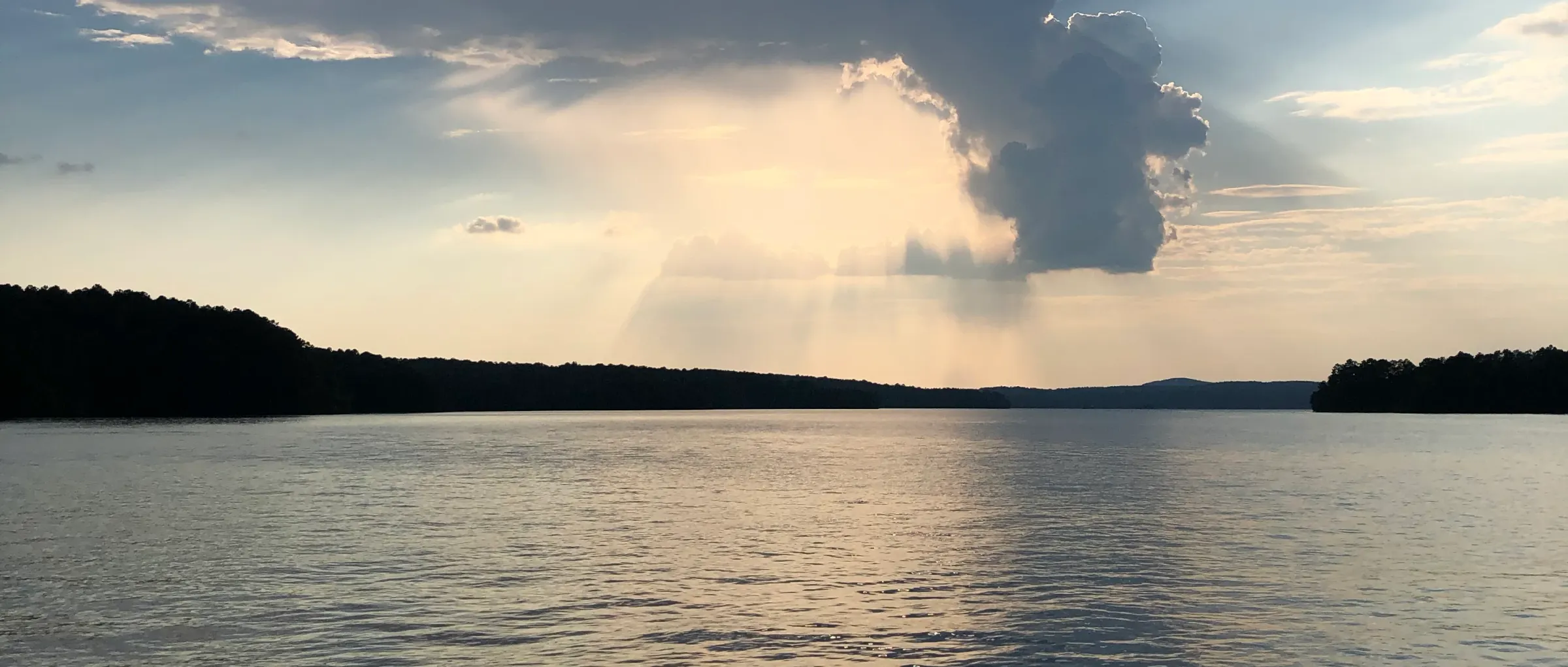 Golden-hour sun rays breaking through clouds over the calm water and tree-lined shoreline of Lake Wedowee